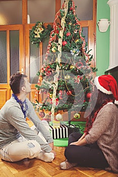 Thoughtful couple sitting under Christmas tree looking at holiday decoration