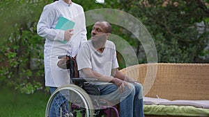 Thoughtful crippled African American man in wheelchair looking away talking to unrecognizable Caucasian caregiver