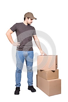 Thoughtful delivery man wearing cap, full length portrait, stands near cardboard parcel boxes, isolated on white background.