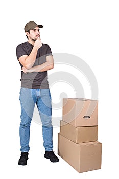 Thoughtful delivery man wearing cap, full length portrait, stands near cardboard parcel boxes, isolated on white background.