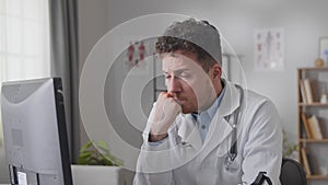 Thoughtful doctor touching chin in medical office. Thinking doctor sitting on workplace front PC while online