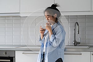 Thoughtful emotionless Asian woman taking medicine and drinking water standing in kitchen near sink