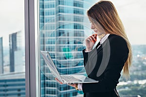 Thoughtful female CEO standing against window in her private office in modern business center holding a laptop reading