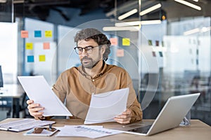 Thoughtful financial advisor in eyeglasses checking and analyzing printed graphs on paper sheets in personal workplace