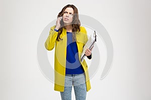 Thoughtful frowning young woman holding clipboard and using smartphone