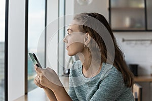 Thoughtful gen Z teen girl holding smartphone, looking out window