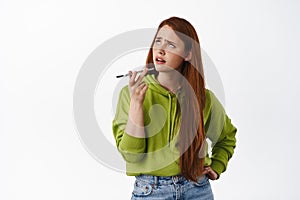 Thoughtful ginger girl talking in speakerphone and thinking, translating words with mobile phone app, sending voice