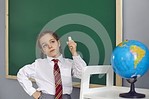 Thoughtful girl with chalk standing in front of blackboard at classroom. Child having difficulty during lesson at school