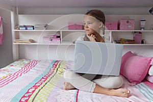 Thoughtful girl looking away while using laptop on bed in a comfortable home