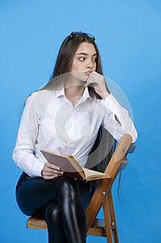 Thoughtful girl with open book, touching lips with hand, while sitting on chair and looking at away.