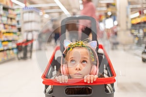 Thoughtful girl wearing headphones in shopping cart at supermarket