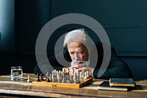 Thoughtful gray-haired senior older man thinking game strategy sitting lying on wooden table with chess board.