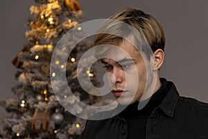 A thoughtful guy 20-25 years old in a black shirt on the background of a decorated Christmas tree.