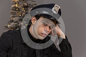 A thoughtful guy of 20-25 years old in a police cap on the background of a decorated Christmas tree.