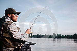 Thoughtful guy is sitting at the edge of water and looking to the right. He holds fly rod in hands. It is evening and