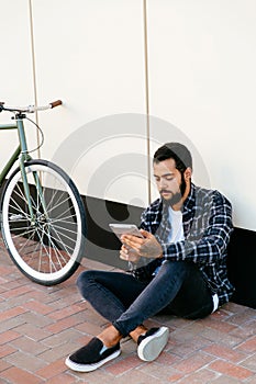 Thoughtful guy using a tablet while sitting near the bike, outdoors