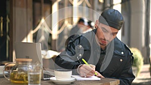 Thoughtful handsome gay man sitting in sidewalk cafe writing letter with pen. Portrait of confident Caucasian LGBTQ