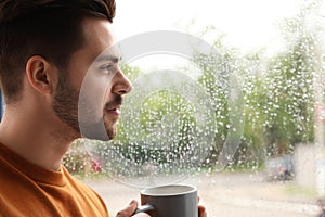 Thoughtful handsome man with cup  coffee near window indoors on rainy day