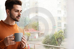 Thoughtful handsome man with cup of coffee near window on rainy day
