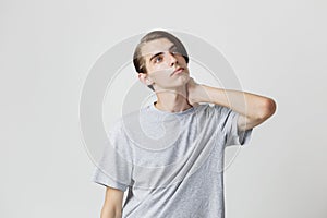 Thoughtful handsome young thin dark-haired guy with blue eyes wearing gray t-shirt standing against light grey