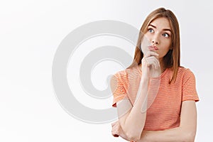 Thoughtful intrigued young fair-haired girl in striped t-shirt, touch chin and pouting curious, look upper left corner