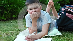 Thoughtful kid boy laying on grass speaking, reading out loud a book. male child writing in exercise book making homework on