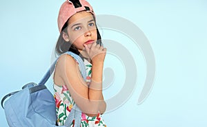 Thoughtful kid wearing a colorful dress, pink cap looking at one side posing over light blue studio background. A pensive little