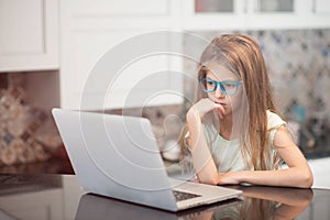 Thoughtful little caucasian girl with long hair wearing glasses sitting in bright kitchen looking on laptop
