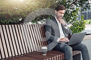 Thoughtful male businessman working on laptop at lunchtime sitting on bench near office