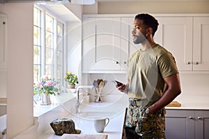 Thoughtful Male Soldier In Uniform With Mobile Phone In Kitchen On Home Leave Looking Out Of Window