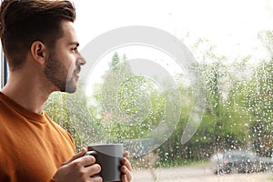 Thoughtful  man with cup of coffee near window indoors on rainy day