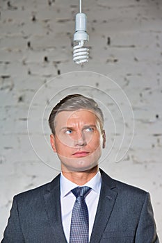 Thoughtful mature businessman looking at energy efficient lightbulb hanging against brick wall at office