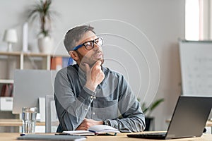 Thoughtful mature man sitting in office and looking aside, working with laptop, looking for creative ideas