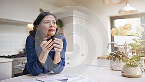 Thoughtful Mature Woman Reviewing Domestic Finances And Paperwork Drinking Coffee In Kitchen At Home