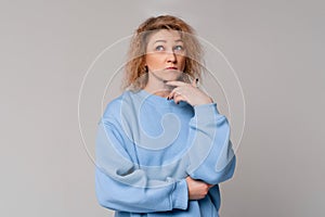 Thoughtful middle age woman with curly blonde hair remembering something, standing with doubtful expression in trendy blue