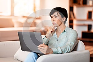 Thoughtful middle aged European woman with laptop in living room