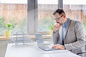Thoughtful middle aged handsome businessman in shirt working on laptop computer in office