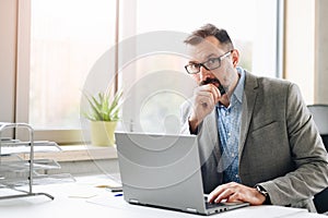 Thoughtful middle aged handsome businessman in shirt working on laptop computer in office
