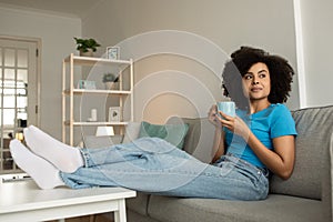 Thoughtful millennial african american curly lady enjoy relax and cup of tea on sofa, think, look at free space