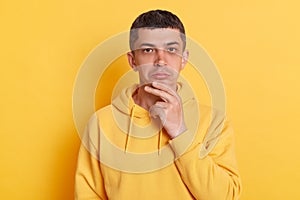 Thoughtful pensive man wearing casual hoodie holding chin looking at camera planning pondering posing isolated over yellow