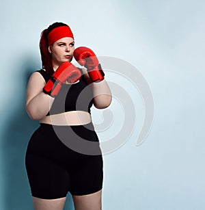 Thoughtful plump  girl in black sporty top, shorts and red boxing gloves looks up thinking over strategy in forthcoming fight.