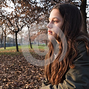 Portrait of a young girl with long hair, sitting at the park, staring far away daydreaming