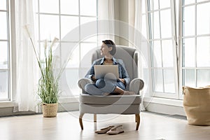 Thoughtful pretty middle aged lady relaxing with laptop in armchair
