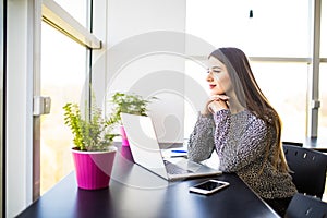 Thoughtful pretty office woman sitting at her desk with laptop, looking into distance with hand on her chin.