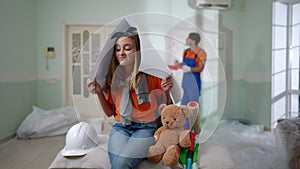 Thoughtful pretty teenage girl with teddy bear sitting indoors covering head with paper as blurred man painting wall