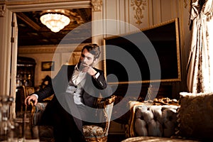 Thoughtful prosperous male involved in business, dressed in formal suit, sits in royal room on comfortable chair, waits photo