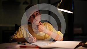 Thoughtful pupil boy studying at home writing in notebook with pen sitting at table under light of lamp.