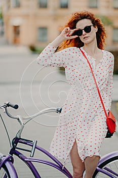 Thoughtful red haired woman focused into distance, keeps hand on frame of sunglasses, poses near bicycle takes break after riding