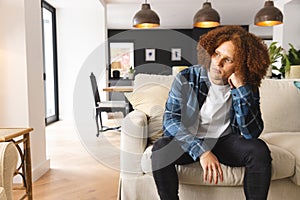 Thoughtful, sad biracial man sitting on couch in living room, looking away, copy space