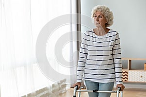 Thoughtful sad injured old woman standing alone using walking frame